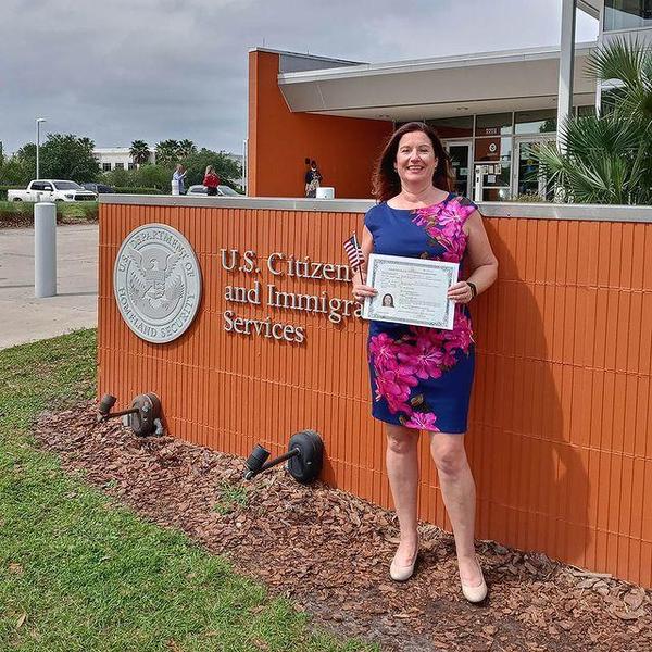 A white woman in a blue and pink floral dress stands outside a U.S. Citizenship and Immigration Services building holding a certificate.