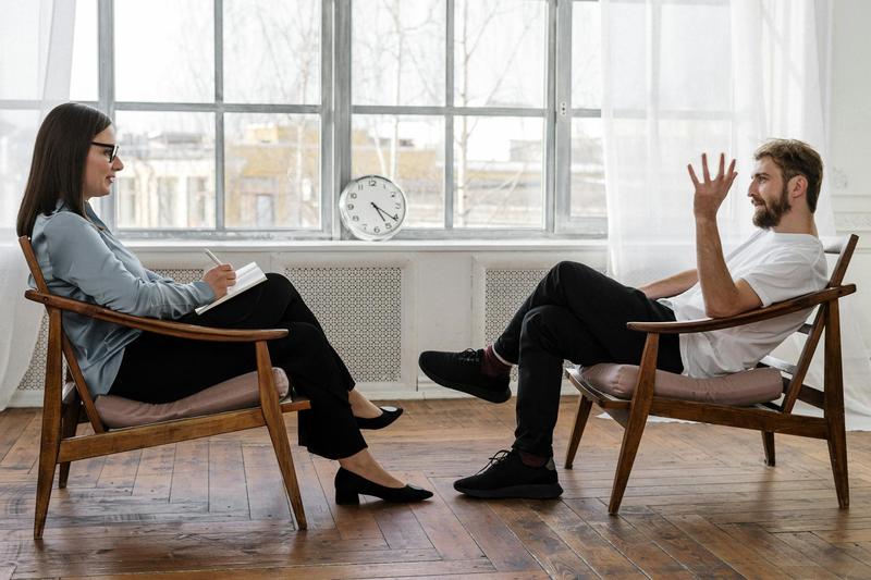A man in a white shirt talks to a woman therapist as they sit in midcentury modern chairs in a minimal room..