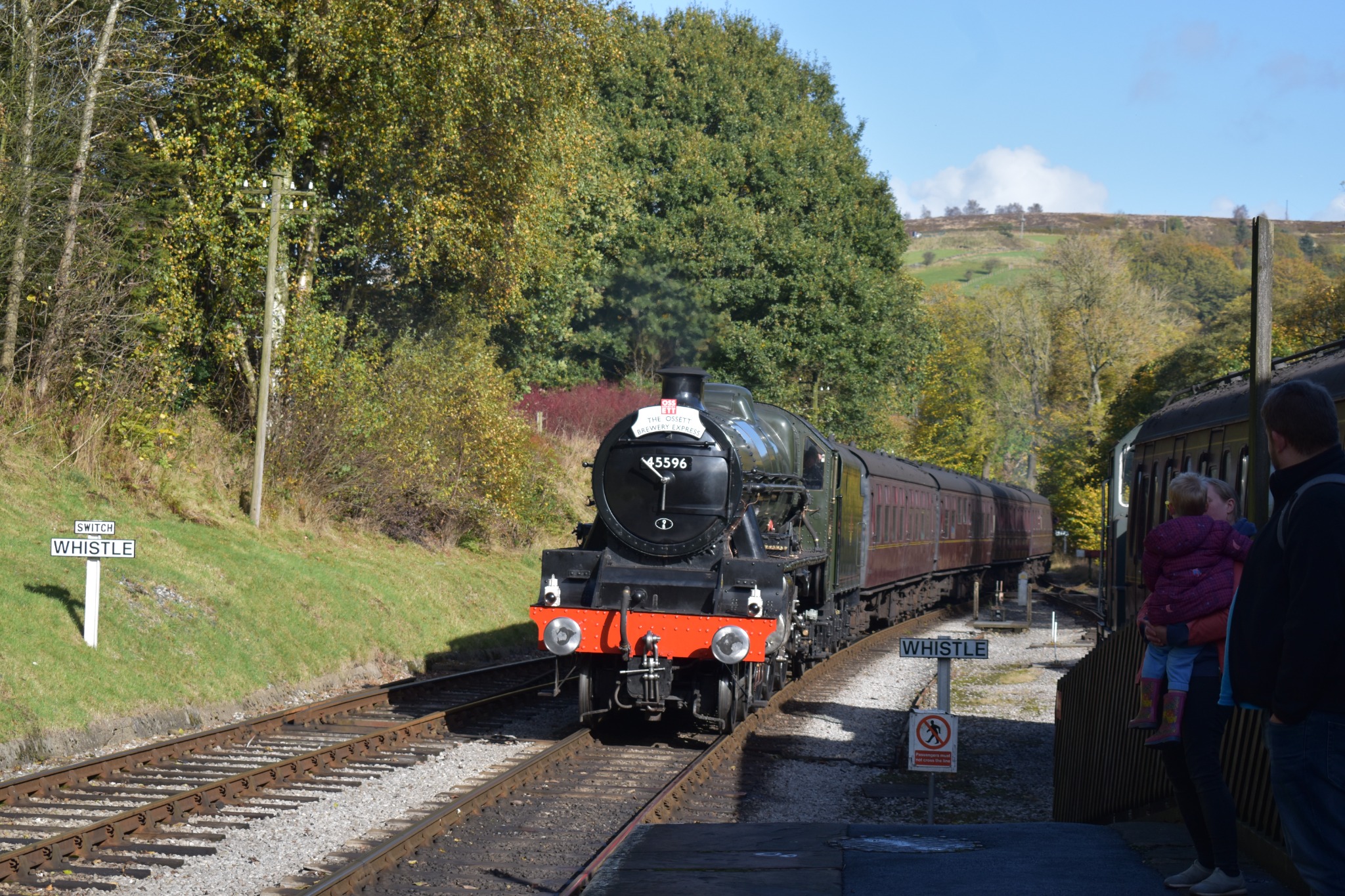 A steam train coming into Oxenhope station. There is a lot greenery on the left side of the tracks and it is a clear day with blue skies. On the end of the platform stands a woman carrying a young child and a gentleman beside them, watching the train come in