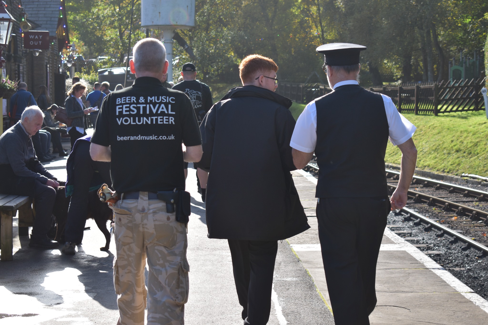 An image of three festival workers/volunteers walking down the platform at Oxenhope station with their backs to the camera. On the left is a gentleman wearing a "beer and music festival volunteer" t-shirt. In the middle is a young man wearing a black coat who works on the trains. On the right is the Station Foreman