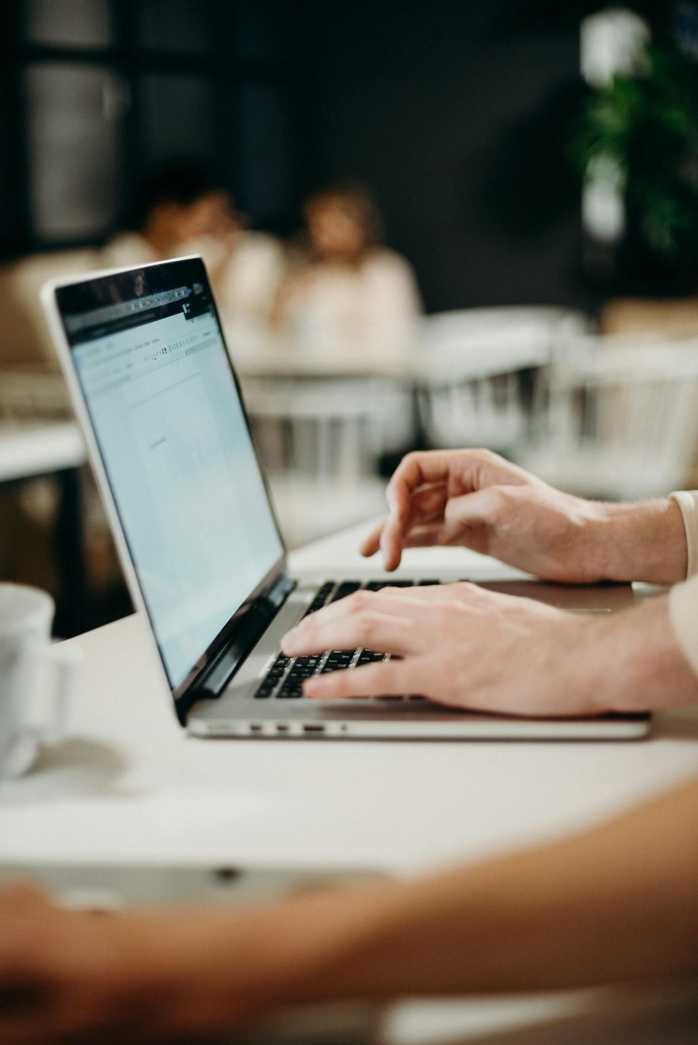 This image shows a side view close up shot of someone's hands typing on a laptop. The background is blurred but the image appears to be taken in a canteen. We can make out a couple of people sat at a table in the background