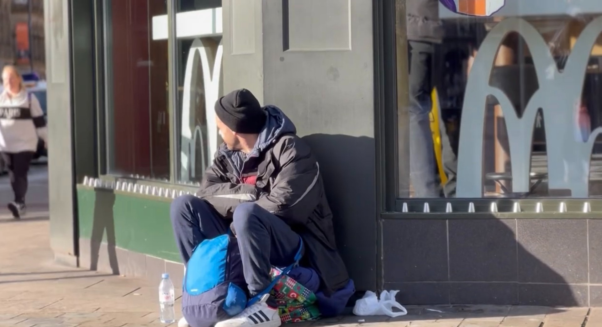 This image depicts a homeless man crouching on the ground outside McDonald's. He's looking away from the camera into the distance. He's wearing a black wooly hat, and has a blue backpack by his feet