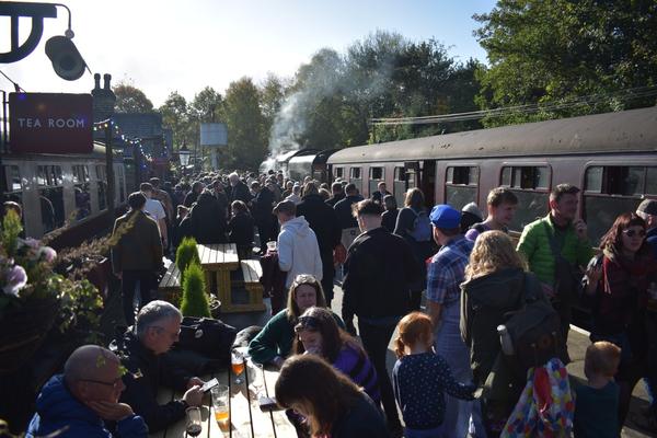 Crowds on people getting off an old steam train at Oxenhope station. There are people sat at a picnic bench on the left. The sun is shining and the sky is blue