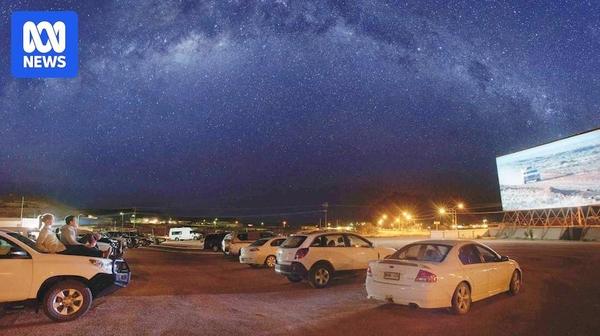 A photo of a drive-in cinema with cars parked facing toward a screen showing a film under a star-filled sky