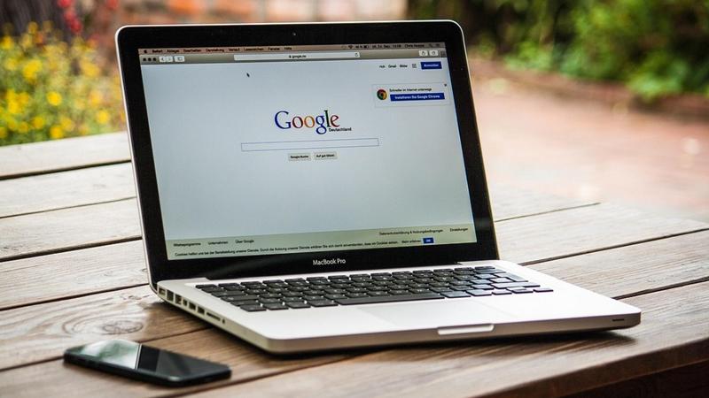 A photo of a Macbook on a wooden bench, with the screen showing the Google homepage.