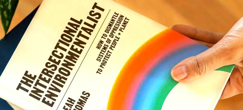 A hand holding a white book with a rainbow on it and the title: "The Intersectional Environmentalist"