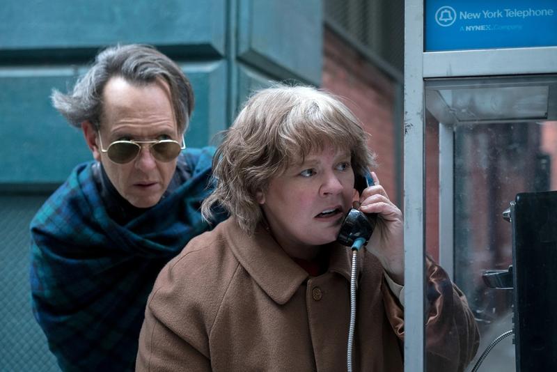 A movie still from Can You Ever Forgive Me? showing a white man (Richard E. Grant) standing behind a white woman (Melissa McCarthy) at a phone booth