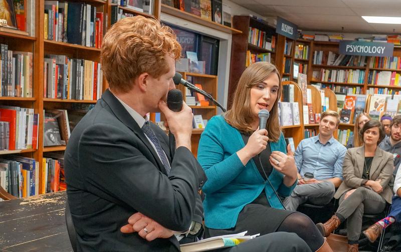A white man with ginger hair sits in a bookstore next a white woman in a teal blazer. They both hold microphones.