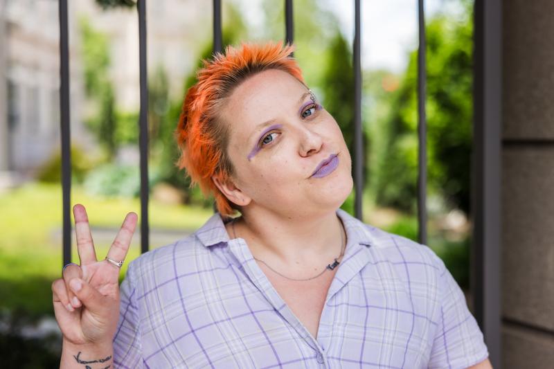 A white person with short orange hair stands in front of an iron gate. Their head is tilted and they have a peace sign up with one hand. They're wearing a purple shirt, purple lipstick, and purple eyeliner. A white person with short orange hair stands in front of an iron gate. Their head is tilted and they have a peace sign up with one hand. They're wearing a purple shirt, purple lipstick, and purple eyeliner.