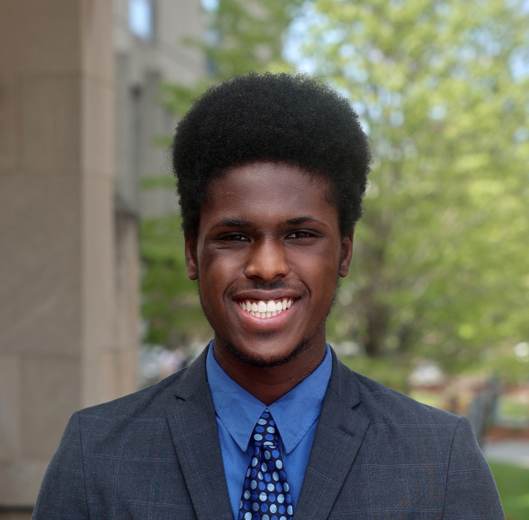 Chinanu, a Black man, smiles in a suit outside. The suit is a plaid black blazer, a baby blue button-down, and a blue polka dot shirt.