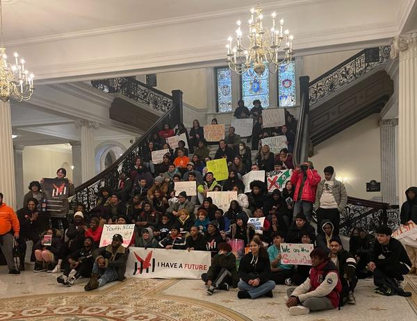 Youth activists stand & sit on a staircase in State House with posters and banners.