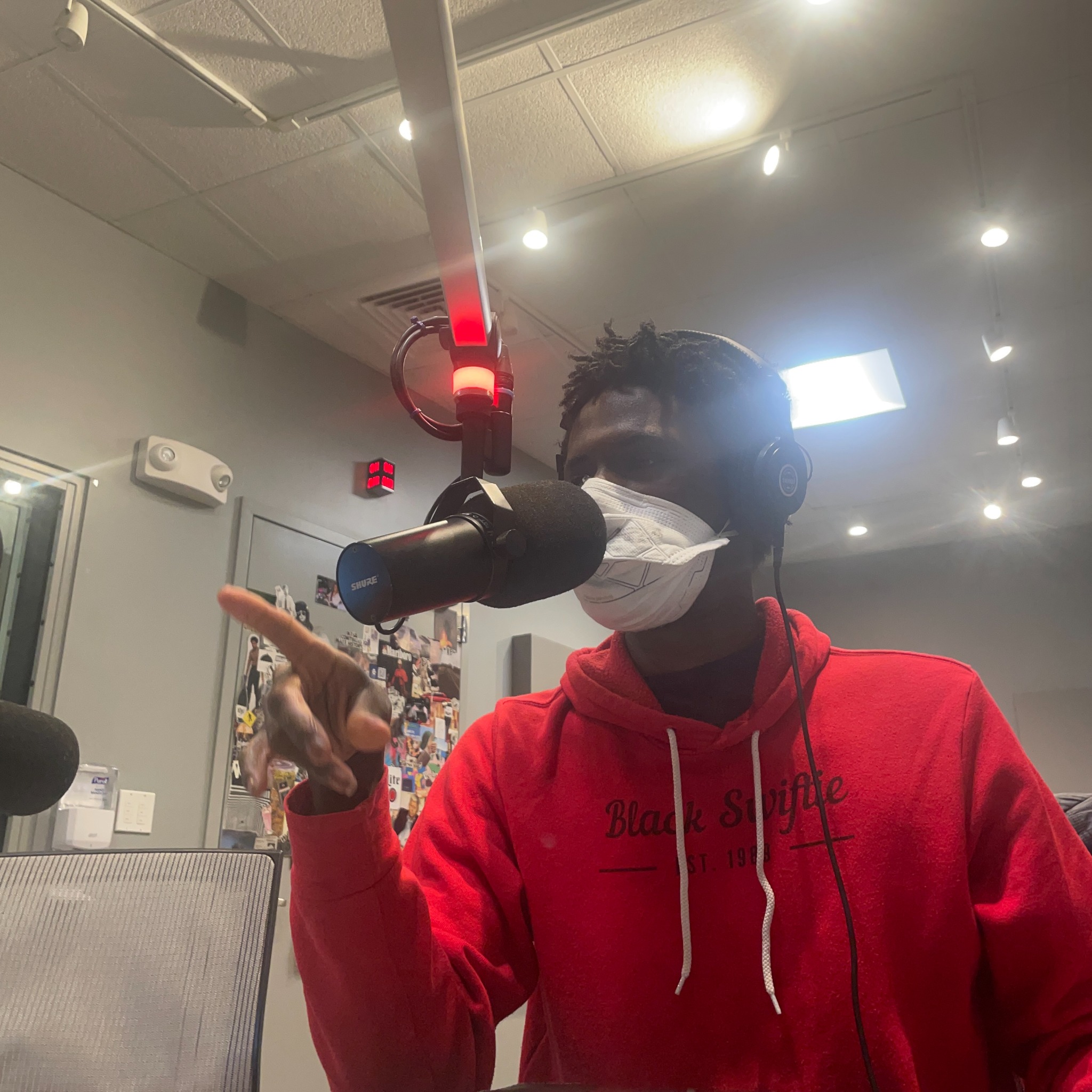 Chinanu points his finger during a live radio broadcast. He sits in a radio studio in a red hoodie, headphones, & a white N95 mask.