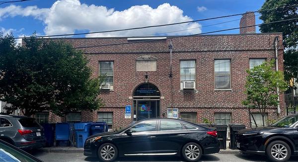 A rectangular brick building on a narrow street with cars parked on either side. There are trees, waste bins, & power lines in front of the building. It’s a partly cloudy day.