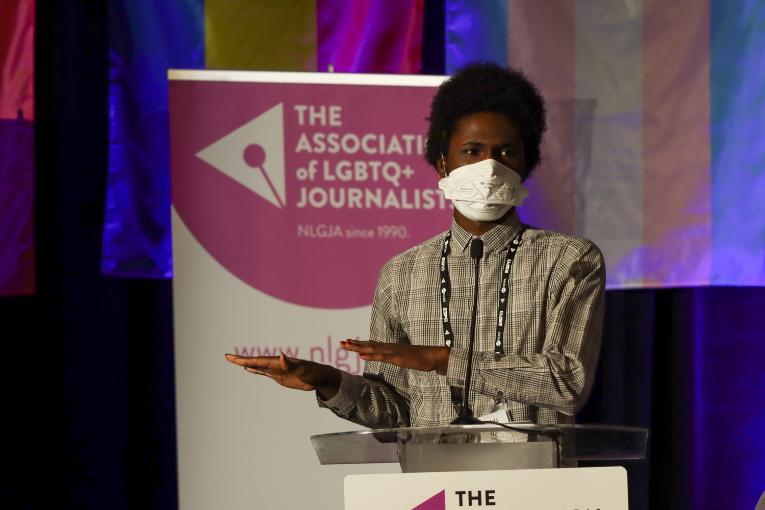 Chinanu presents on stage at the NLGJA convention. He wears a white & black plaid shirt, a lanyard, a white N95 mask, & red nails. He stands in front of various pride flags.