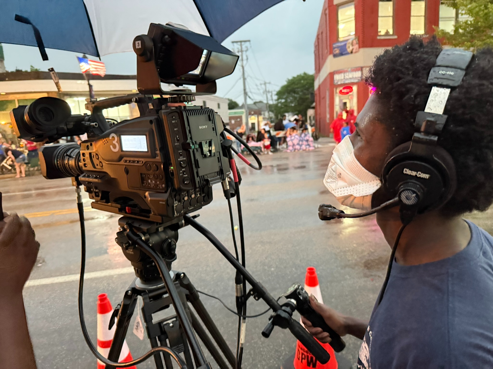 Chinanu operates a camera in the rain during a parade. Someone else holds an umbrella over the camera in front of him.