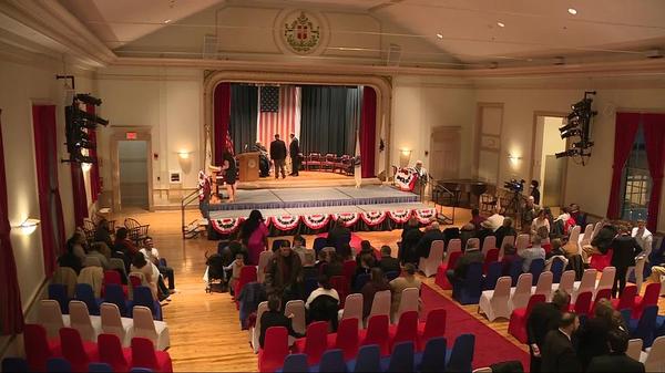 Inauguration hall. A few people stand on stage arranging items as dozens of people sit in red, blue, & white chairs on the hall floor.