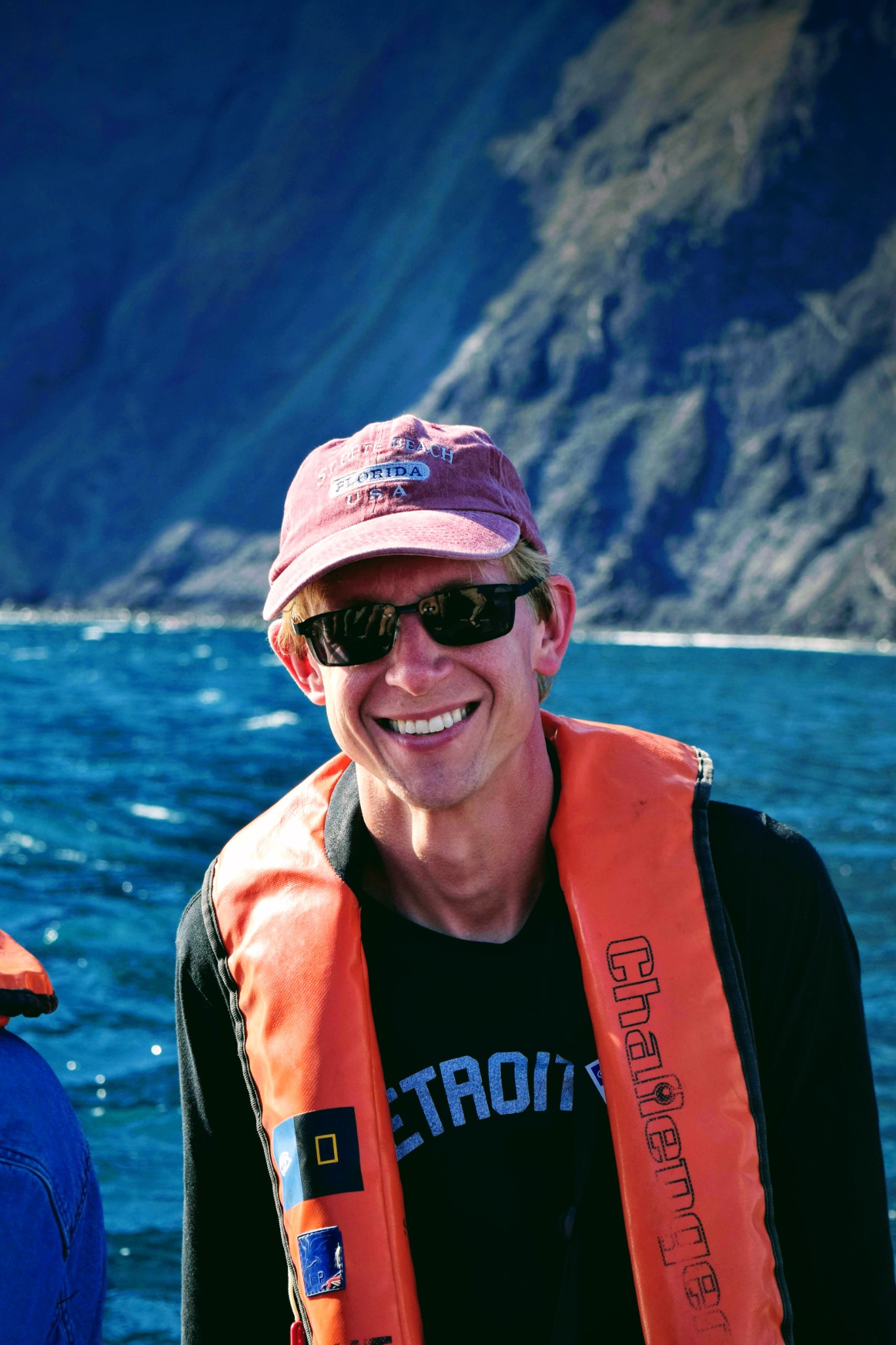 A man in an orange life jacket smiles on the water with a tall mountain behind him.