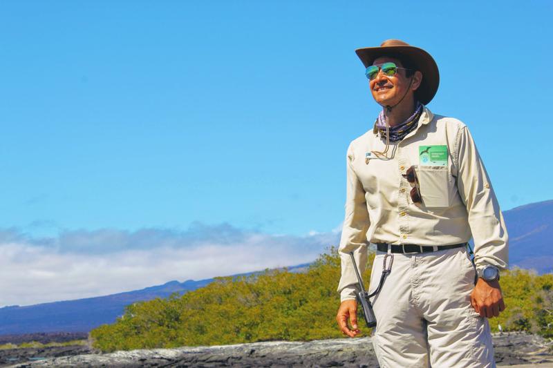 Galapagos Islands naturalist Celso Montalvo, smiling and wearing a tan shirt, pants, and wide-brimmed hat, standing on Isabela Island in front of a mountain.