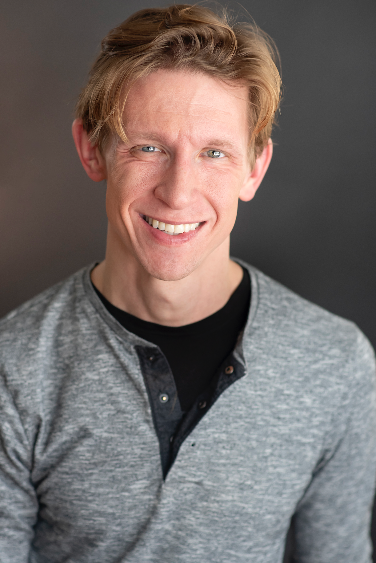 A professional headshot for a man with short blonde hair, a grey shirt, and black undershirt. He is smiling.