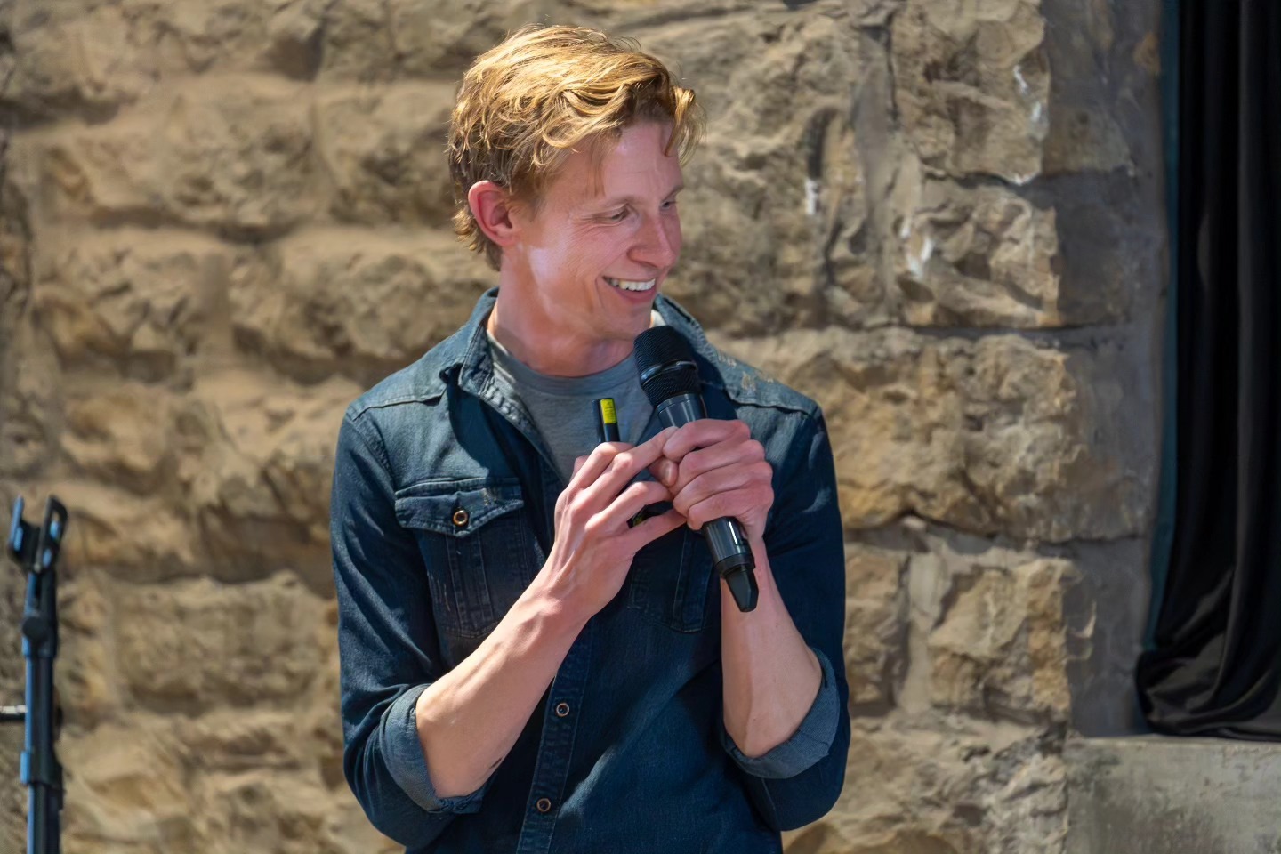 A man with blonde hair in a blue denim shirt smiles while holding a microphone and a small clicker