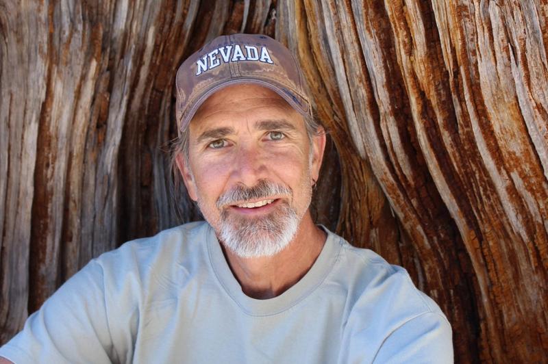 A man with a grey goatee and Nevada hat smiles in front of a giant tree