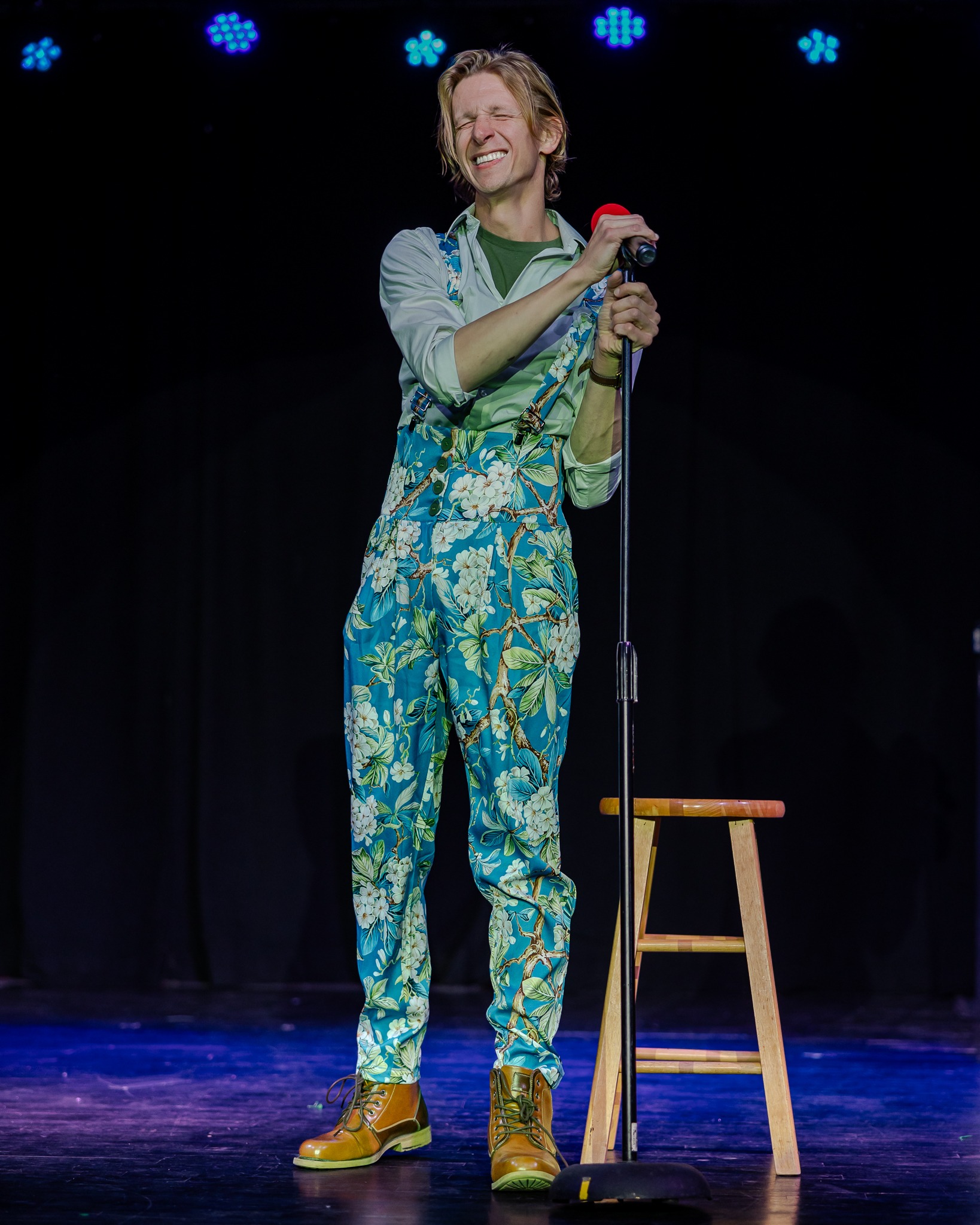 A blonde man on a dark stage smiles while holding a red microphone. He is wearing turquoise overalls with white flowers on them.
