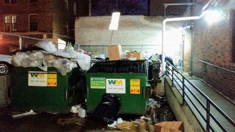 Two dumpsters overloaded with trash in a lit alley at night