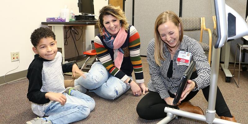 Two adults and a child sit on the floor looking at an assistive device with a screen. The adult on the right is laughing.