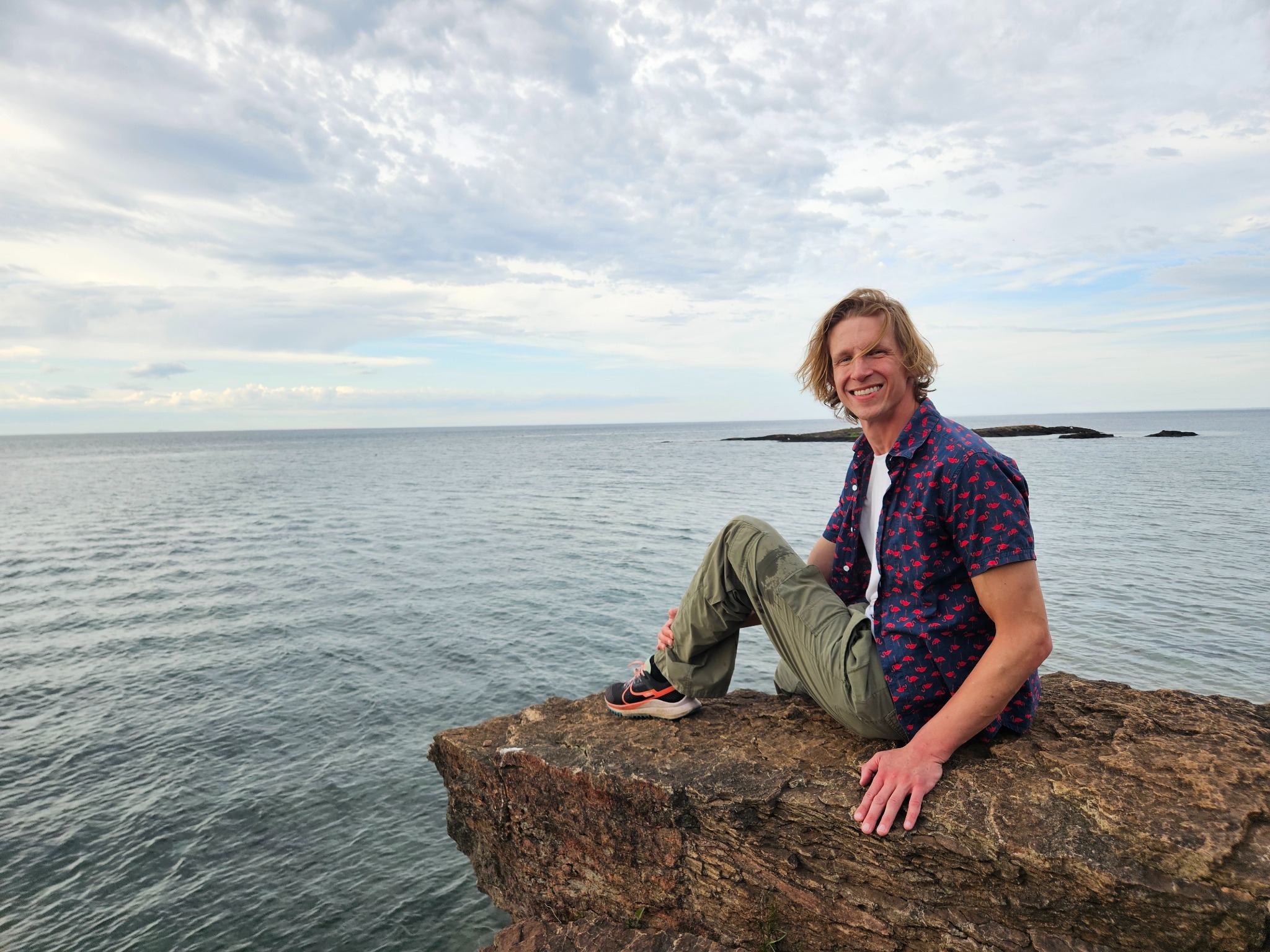 A blonde man in green pants and a blue shirt sits on a cliff edge looking over a large body of water