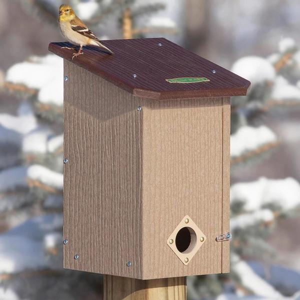 This product photo shows a winter bird house with the round entrance on the bottom and a goldfinch perched on top (in its winter plumage). There is snow on the trees behind the bird house and the bird house itself is made from light and dark brown plastic.