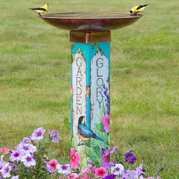 This photo of a colorful bird bath has two goldfinches drinking water from the wide copper basin on top of the square post. The decorative post says, "Garden" and "Glory" along two sides, accented with petunias and birds and more.