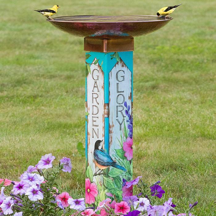 This photo of a colorful bird bath has two goldfinches drinking water from the wide copper basin on top of the square post. The decorative post says, "Garden" and "Glory" along two sides, accented with petunias and birds and more.