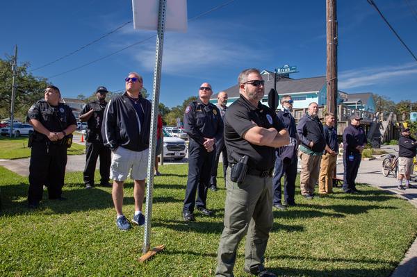 A group of police officers and city personnel stand outdoors on a sunny day, listening during a Veterans Day event near a street corner.