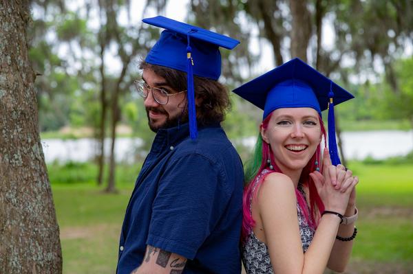 Two graduates wearing blue caps stand back-to-back outdoors, smiling playfully at the camera.