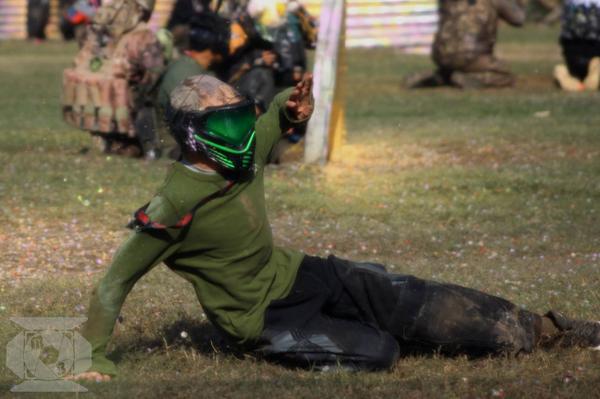 A person wearing a green shirt, padded pants, and a green paintball mask slides on the ground during a paintball game, with other players crouched in the background.