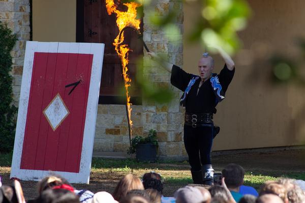 A performer at the Texas Renaissance Festival holds a flaming whip while balancing a small object on his head in front of a cheering audience.