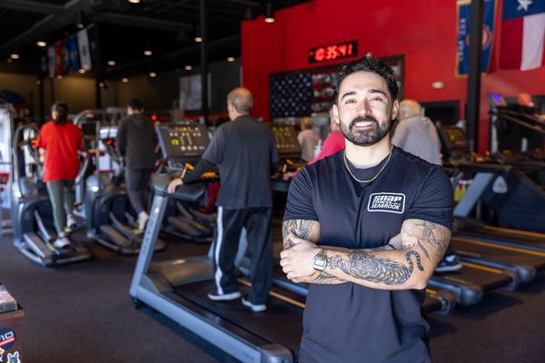 Christopher Garcia, owner of Snap Fitness Seabrook stands smiling in front of treadmills at a gym where people are exercising.