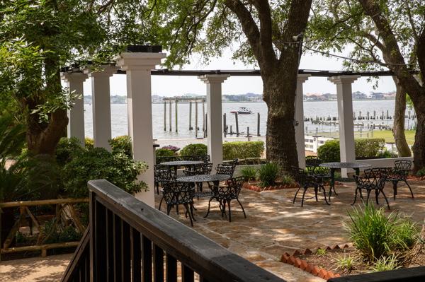 Patio with black metal tables and chairs shaded by trees, overlooking Clear Lake with boats and waterfront homes in the distance.