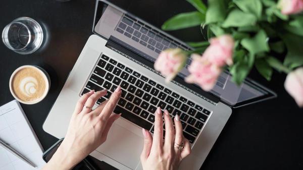 Workspace setup for a bootstrapping founder managing an online business with low cost tools and a virtual assistant, featuring a laptop, planner, coffee, and flowers on a neatly arranged desk.