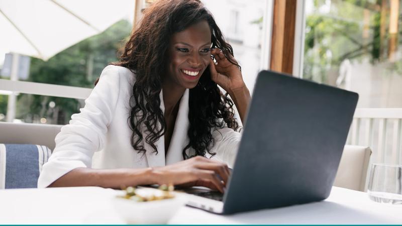 A smiling Black woman entrepreneur working on her laptop at an outdoor cafe, showcasing the freedom of knowing What to Set Up Before Hiring a Virtual Assistant (Beginner Guide).