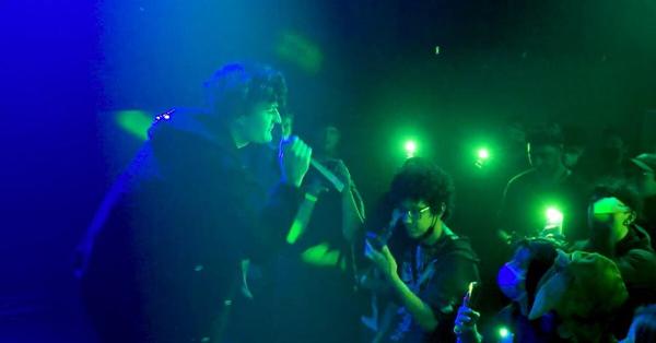 A frame from my documentary of emo rap artist Hamilton performing to a small crowd in an underground venue, surrounded by blue and green lights.