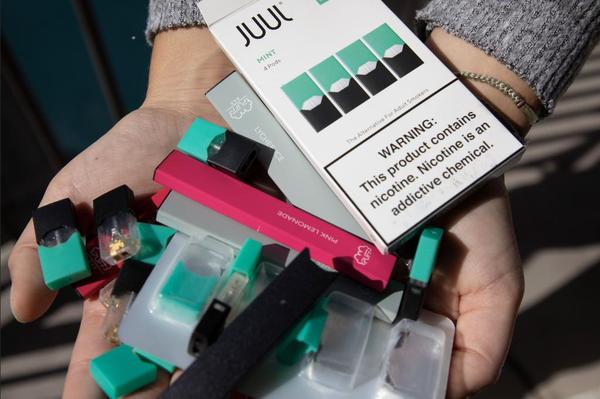 A person holding JUUL vape pods, a JUUL battery, and a Pink Lemonade flavored PUFF Bar vapor device.
