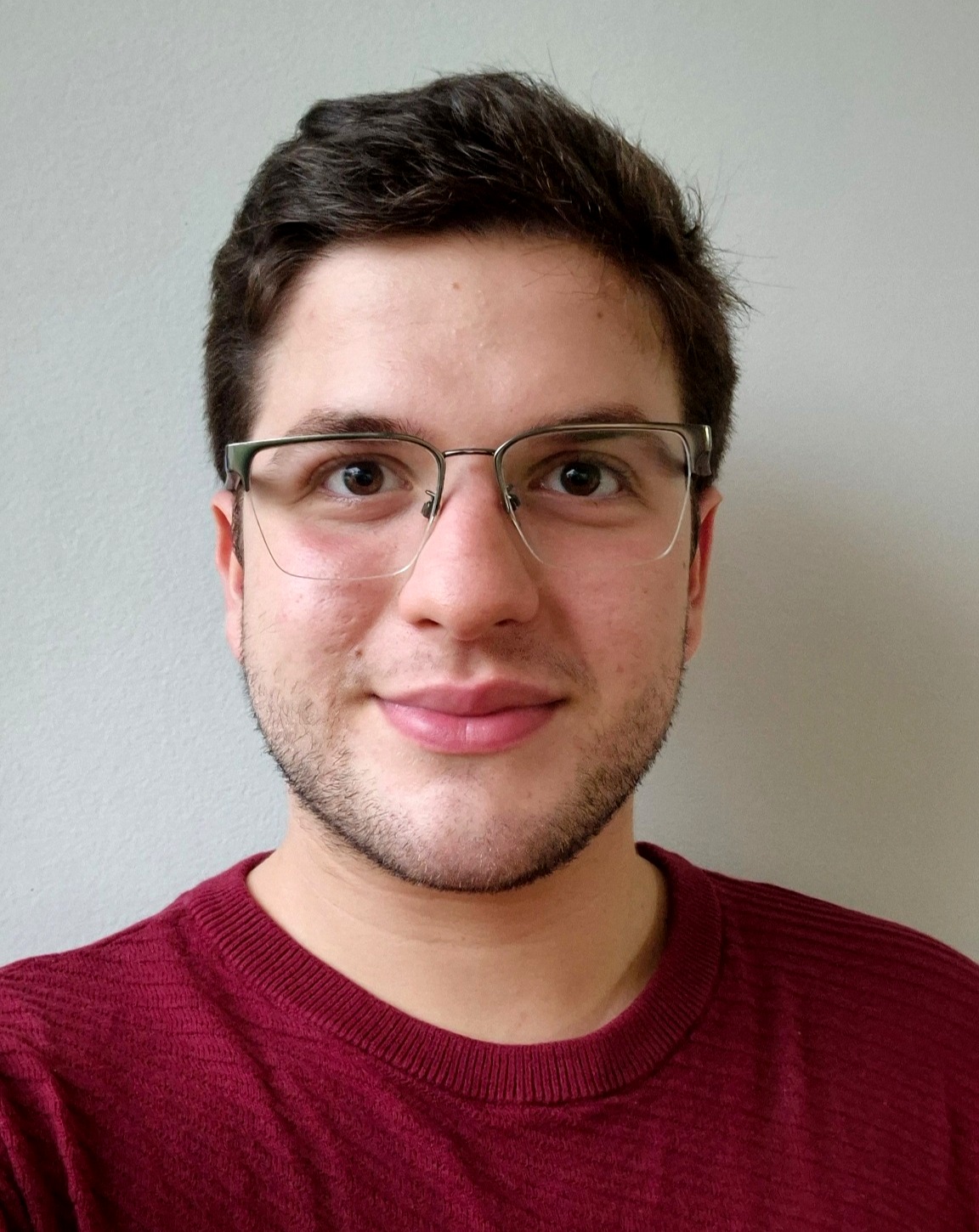 Retrato frontal de um homem jovem, de óculos, com barba curta e cabelo castanho, usando suéter vinho, em fundo neutro claro, olhando diretamente para a câmera com expressão serena e confiante.