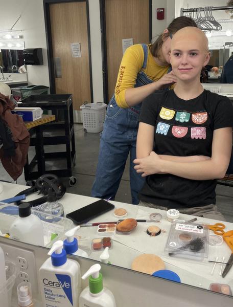 White actress with bald cap smiles at mirror with arms cross as woman in back works on neck