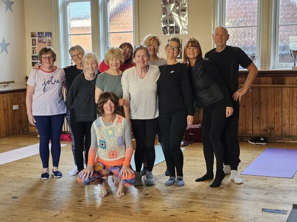 A welcoming mixed group of men and women of all ages posing together at a Daisy Chain Yoga class in a local community hall.