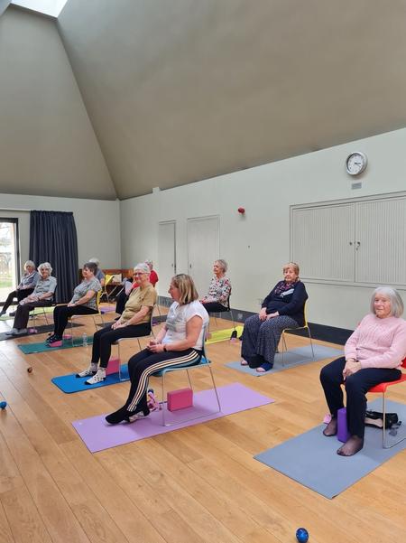 A gentle Chair Yoga class at Daisy Chain Yoga, featuring a group of students practicing seated poses on yoga mats in a bright community hall.