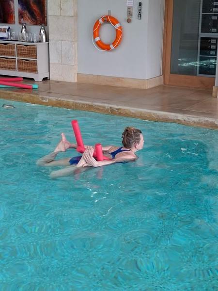 A woman practicing a floating yoga pose in a bright indoor pool, using red foam noodles for buoyancy and support. Daisychainyoga Aqua classes