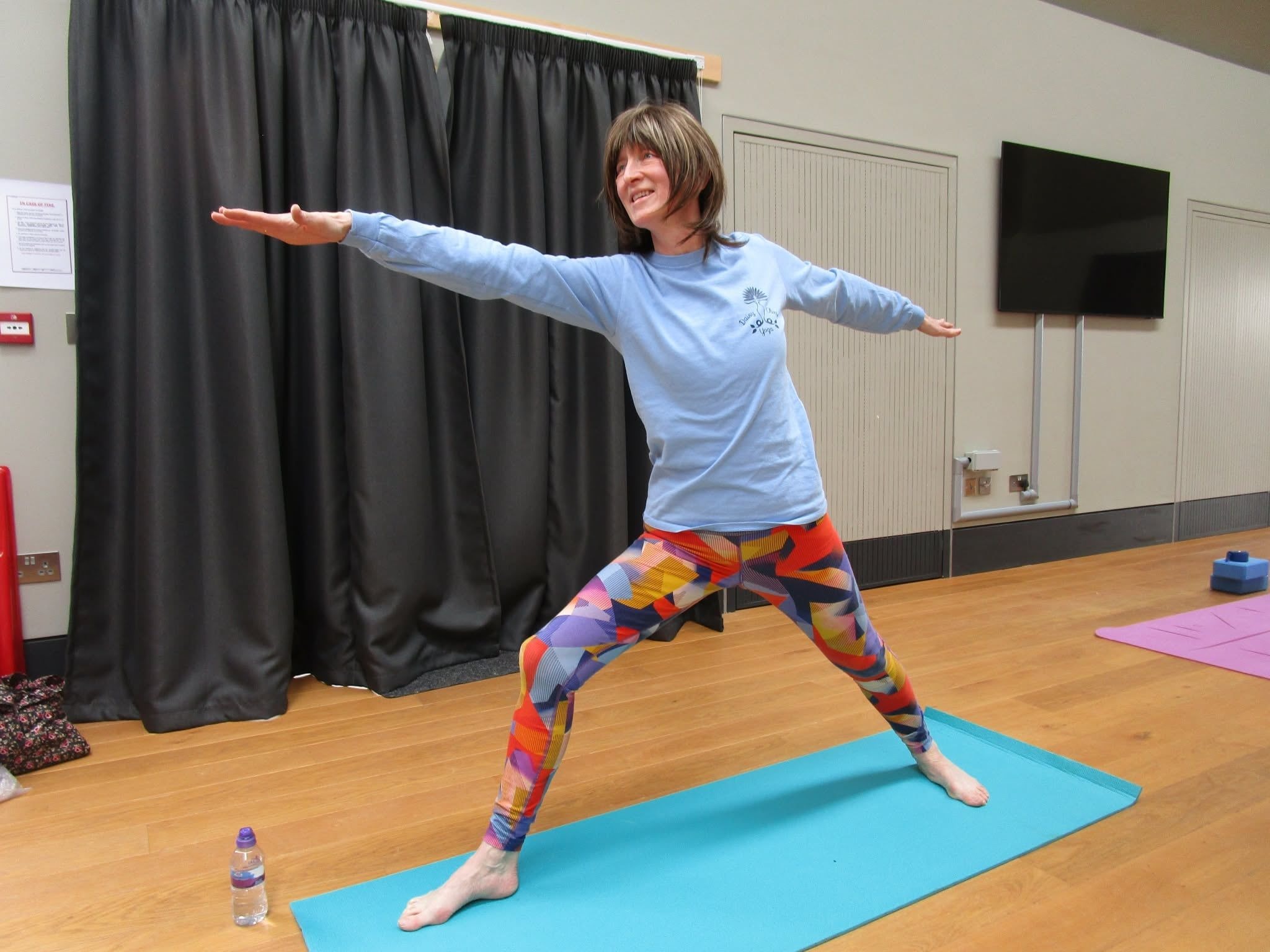 Rachel Metcalfe of Daisy Chain Yoga practicing a Warrior II pose during a yoga class in a community hall.