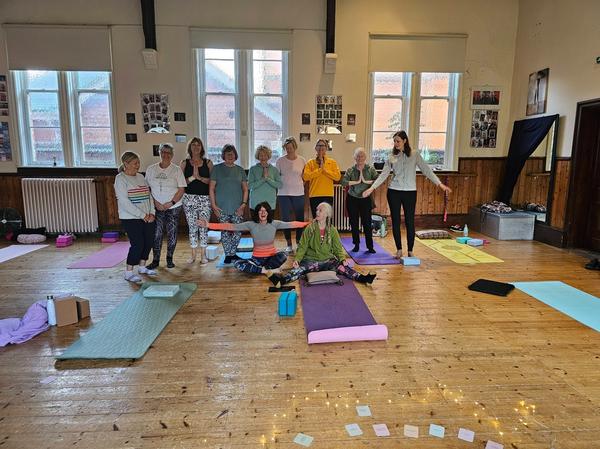 A friendly group of yoga students and instructor Rachel Metcalfe smiling in a village hall class at Daisy Chain Yoga.
