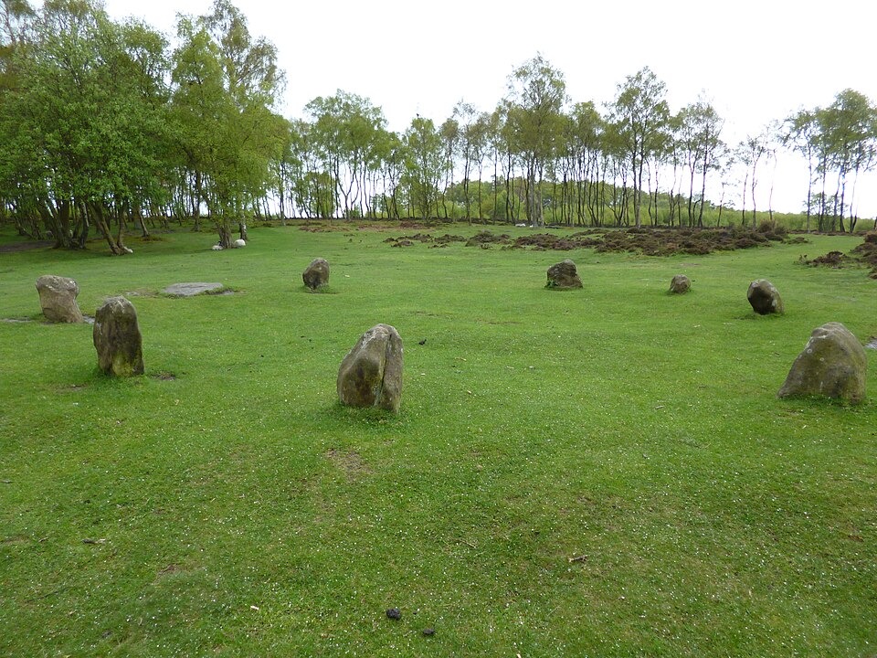 The low standing stones of the Nine Ladies Stone Circle on Stanton Moor in Derbyshire, surrounded by grass and woodland.